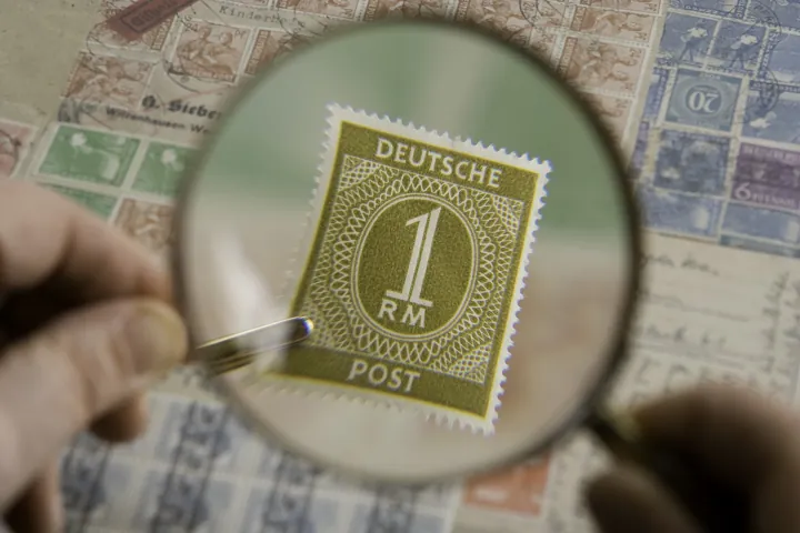Photo d’origine : Une loupe qui grossit un timbre-poste ancien posé sur une planche de timbres, tenue entre les doigts.