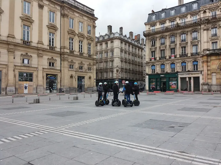 Photo d’origine : Un petit groupe de touristes en gyropodes sur une place de Lyon, devant des immeubles anciens.
