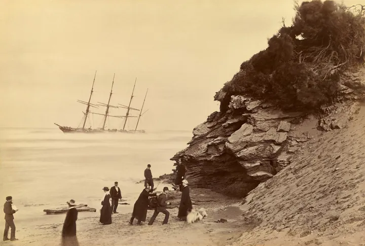 Photo d’origine : Un grand voilier à quatre mâts échoué au large d'une plage, avec quelques promeneurs sur le sable.