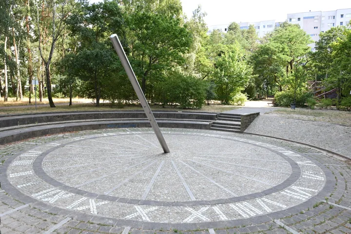 Photo d’origine : Un grand cadran solaire au sol dans un parc, avec son style métallique incliné et ses heures en chiffres romains.